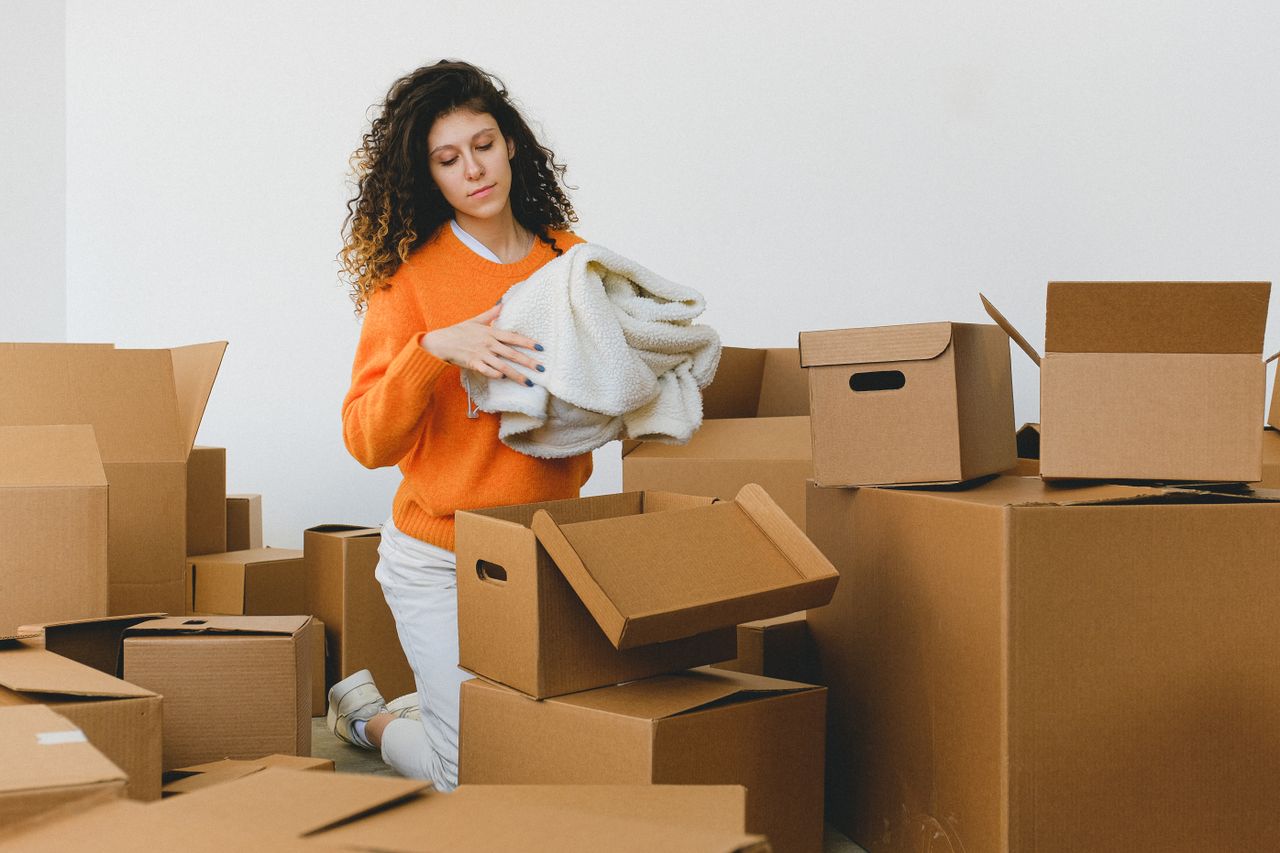 Jeune femme concentrée aux cheveux bouclés, agenouillée au sol et rangeant des vêtements dans des cartons avant de déménager dans un nouvel appartement